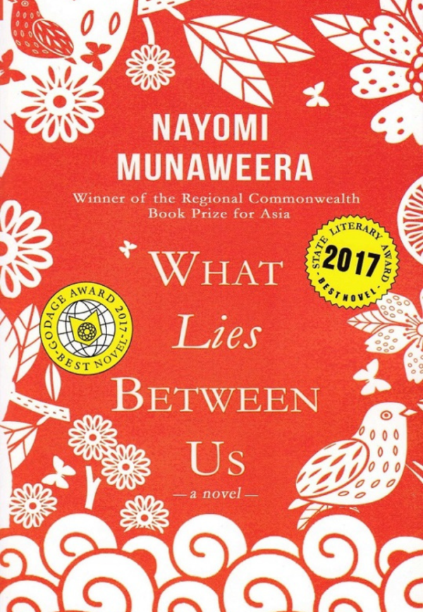 Cover of "What Lies Between Us" by Nayomi Munaweera, featuring a vibrant red background with white floral designs and award stickers.